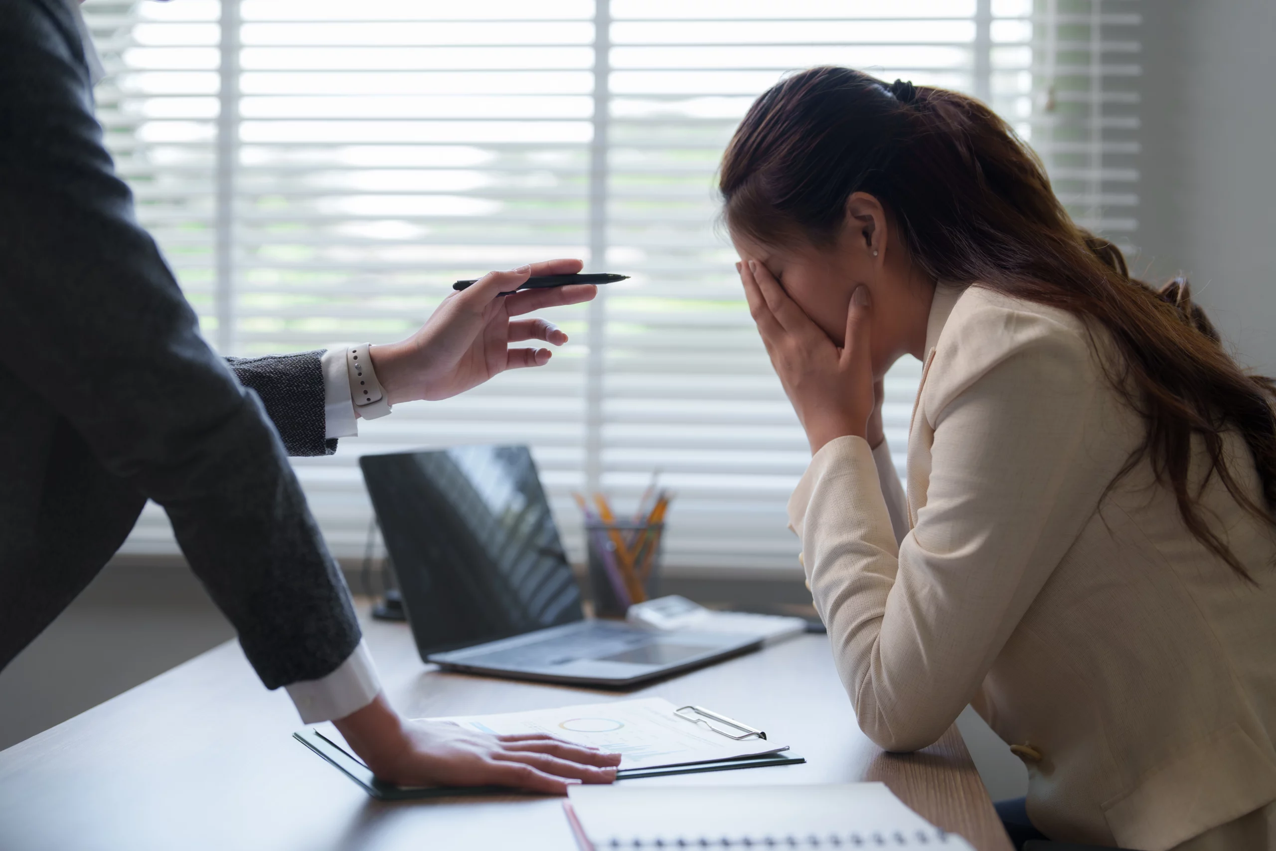 Stressed woman at desk covering her face while colleague points pen, illustrating workplace retaliation and employee rights issues in California.